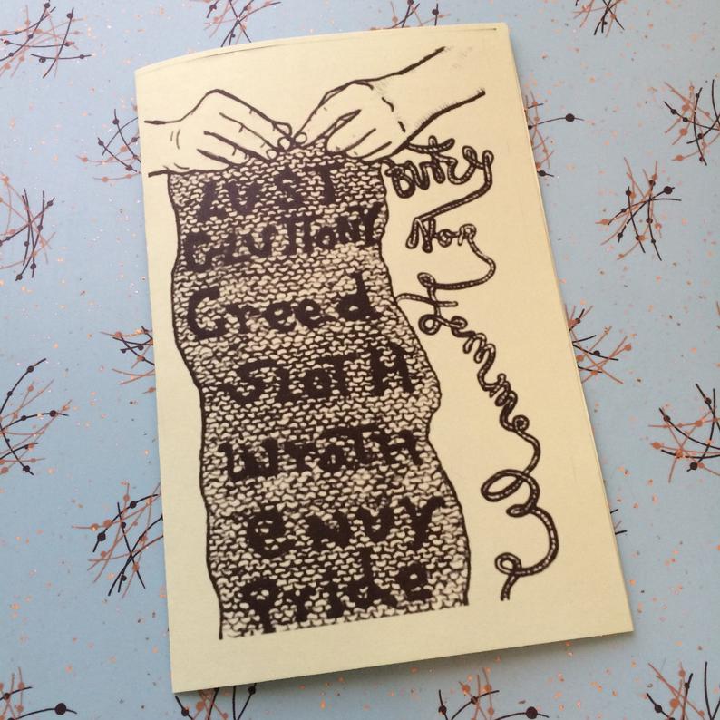 photo of zine on 1950s looking formica. cover is the handwritten title written from bottom to top in cursive. on the left are hands knitting a scarf with the names of the seven deadly sins written on it.