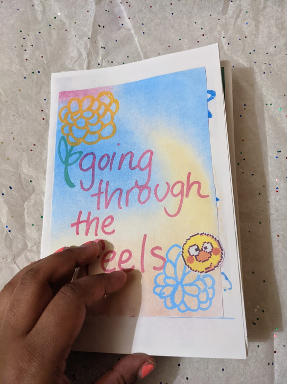 photo of colorful zine cover with a handwritten title, held in the lower left corner by a brown hand