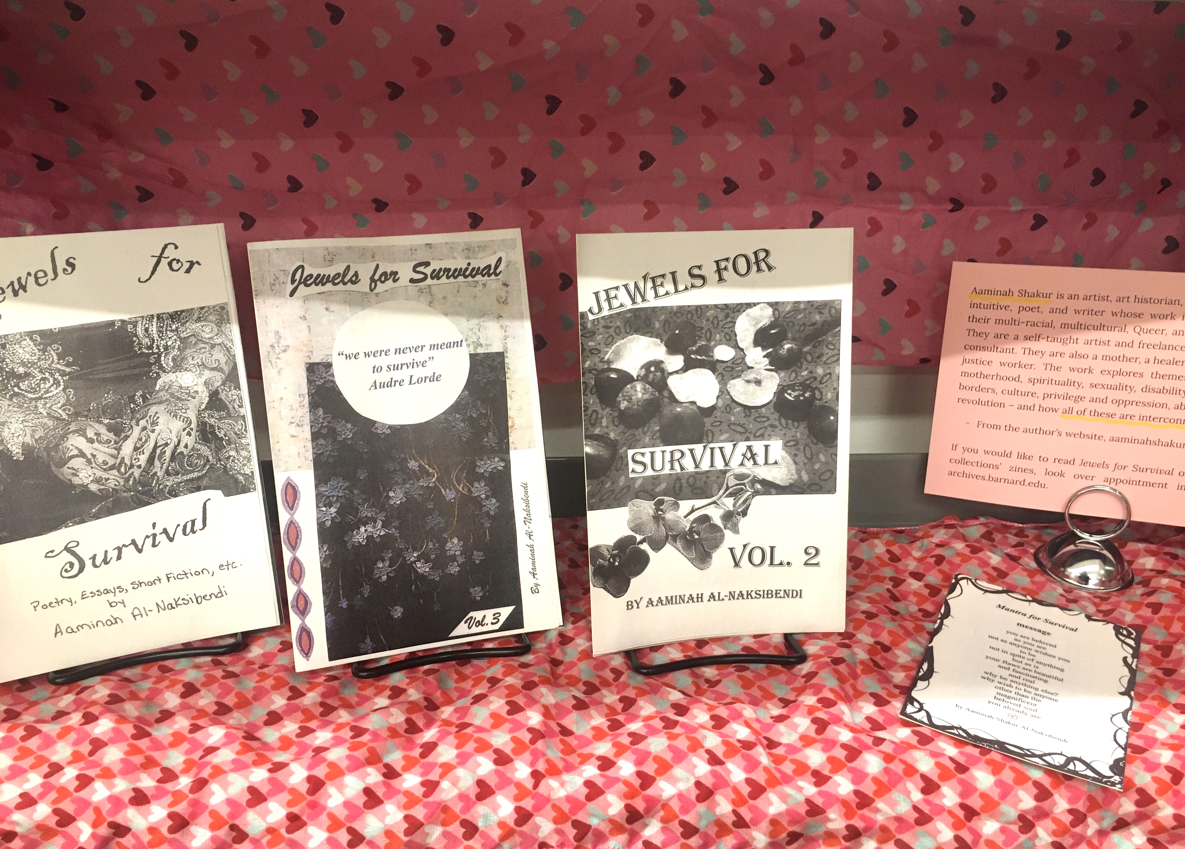 A zine display featuring three photocopied zines, a little pamphlet with a poem written on it by the author, a label for the display, and pink fabric behind the display with colorful hearts