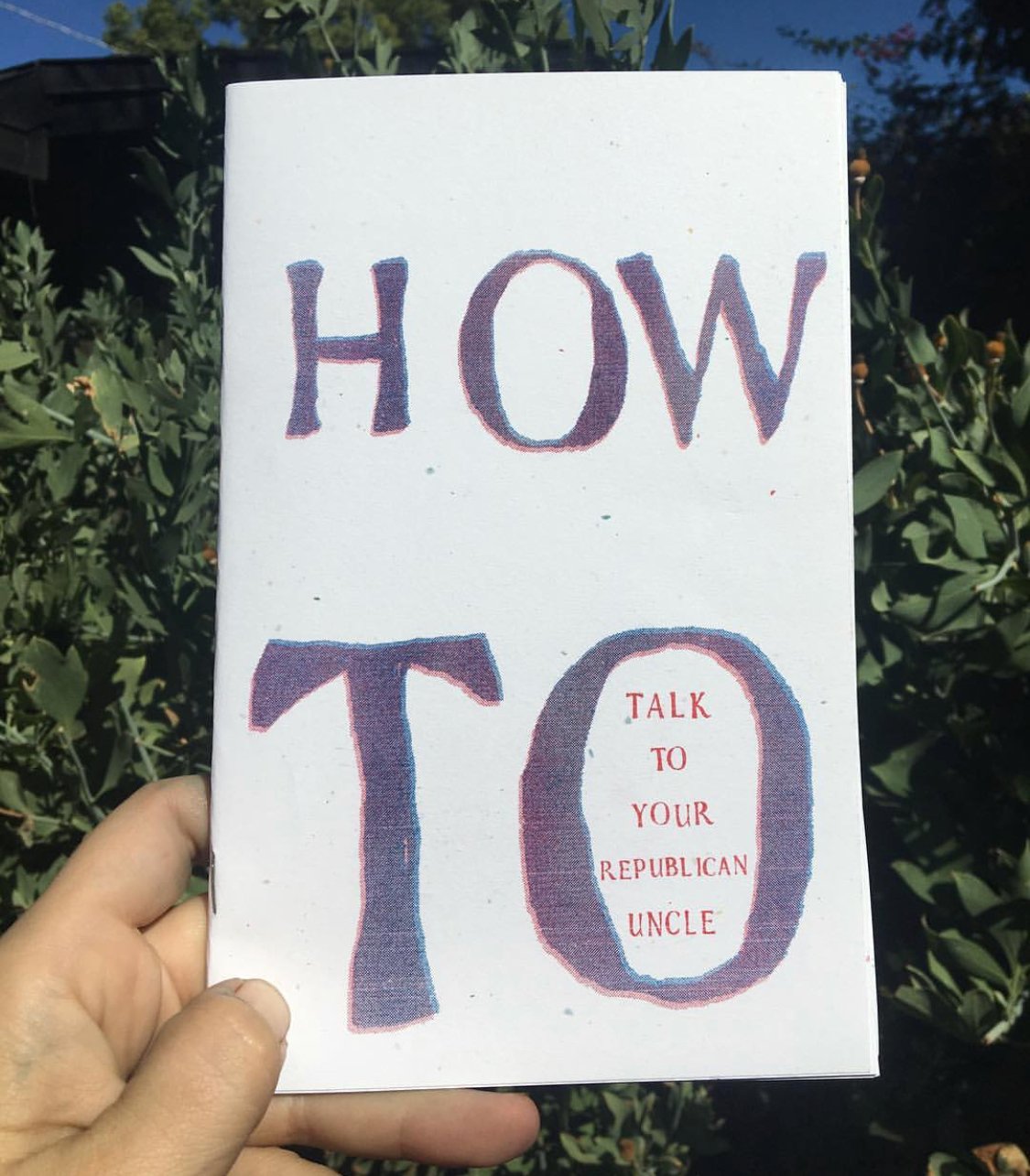 photo of hand holding a color printed zine: "How" and "To" in large letters, "Talk to Your Republican Uncle" in smaller letters inside the "o" in "to"