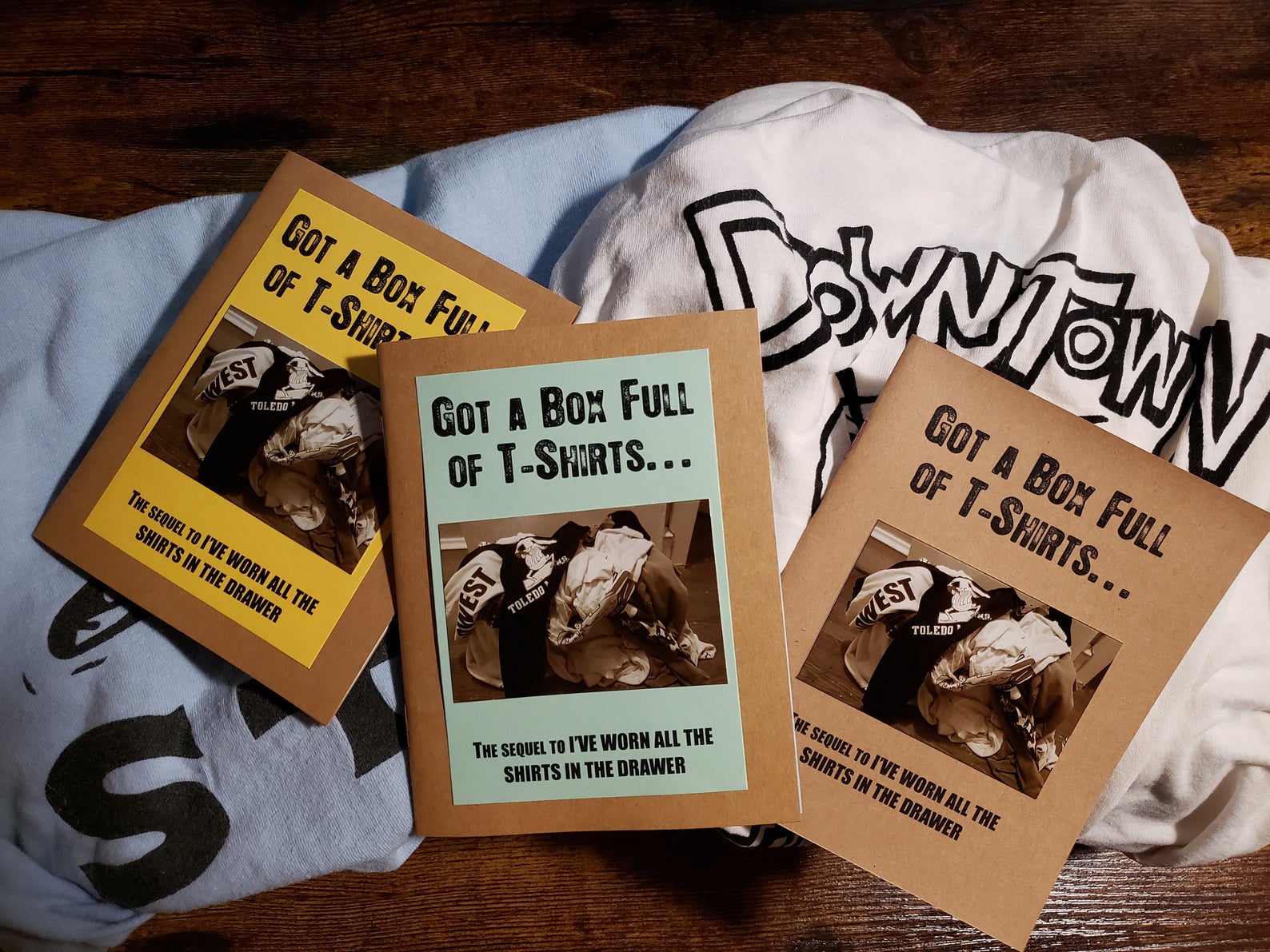 photo of three zines on a pile of t-shirts. zines have brown covers and a glossy photo insert. 